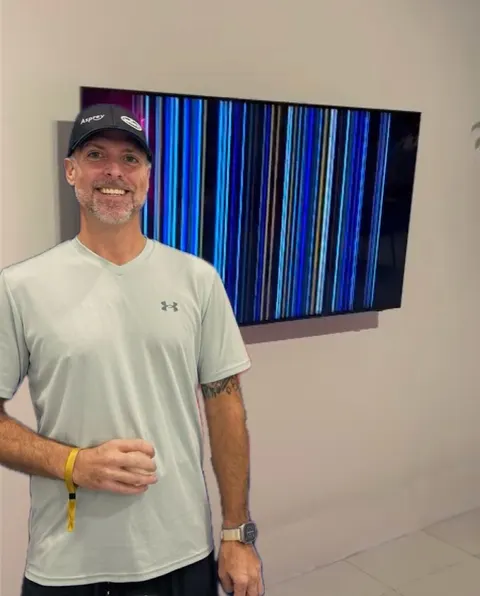 A man in a cap and gray Under Armour shirt smiles for a photo beside a wall-mounted television displaying a Kanetix Light Waves generative piece rendered as vertical blue and white light bars, in a white-walled gallery.