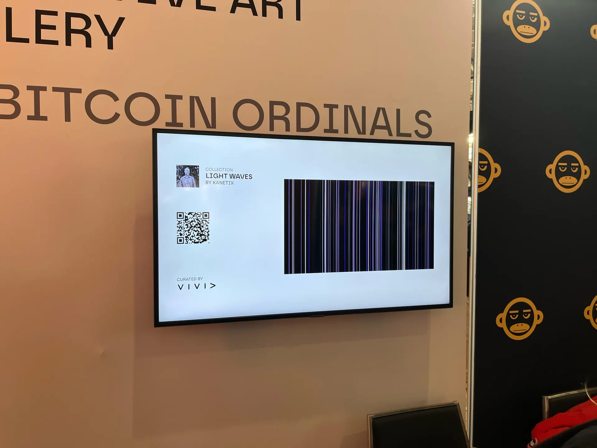 A wall-mounted screen reading "Collection Light Waves by Kanetix, curated by Vivid," showing a QR code beside a Light Waves piece rendered as vertical black and blue light bars, set against a "Bitcoin Ordinals" gallery wall with a repeating monkey-logo backdrop.