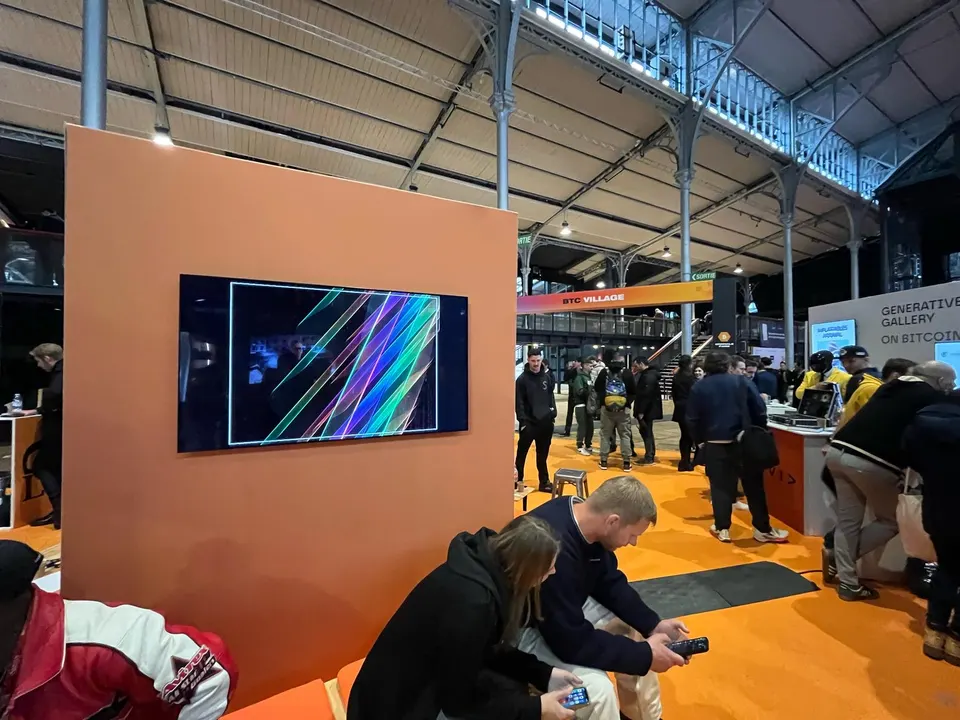 A wide view of the NFT Paris exhibition hall inside a glass-roofed train station, with an orange partition wall displaying a colorful generative artwork on a wall-mounted screen, visitors seated and standing nearby, and a "BTC Village / Generative Gallery on Bitcoin" banner overhead.
